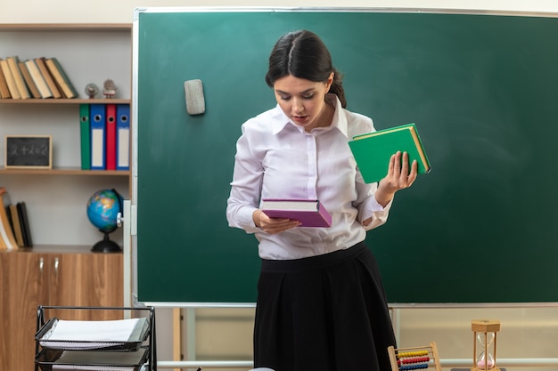 Pensando na jovem professora segurando e olhando para livros em frente ao quadro-negro na sala de aula