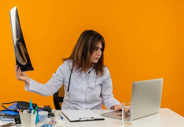 Pensando na médica de meia-idade vestindo túnica médica com estetoscópio sentada na mesa de trabalho no laptop com ferramentas médicas segurando um raio-x e olhando para o laptop na parede laranja isolada