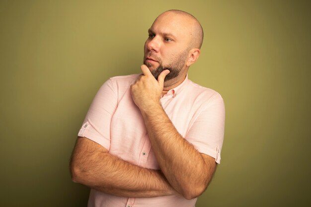 Pensando olhando para o lado de um homem careca de meia-idade vestindo uma camiseta rosa agarrou o queixo isolado na parede verde oliva