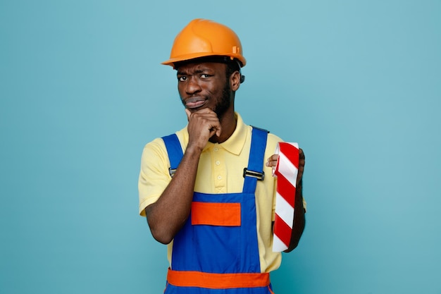 Pensando que agarrou o queixo segurando a fita adesiva jovem construtor americano africano de uniforme isolado em fundo azul