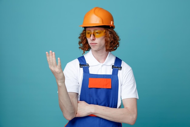 Pensando segurando e olhando para a mão jovem construtor de uniforme isolado em fundo azul