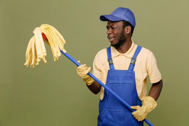 Pensando segurando e olhando para o esfregão jovem limpador afro-americano de uniforme com luvas isoladas em fundo verde