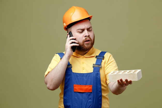 Pensando segurando o tijolo fala no telefone jovem construtor de uniforme isolado em fundo verde