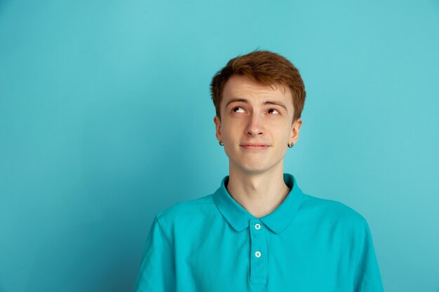 Pensando, sonhando. Retrato moderno de jovem caucasiano isolado na parede azul, monocromático. Lindo modelo masculino. Conceito de emoções humanas, expressão facial, vendas, anúncio, moda.