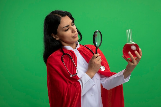 Pensativa jovem super-heroína vestindo túnica médica com estetoscópio segurando e olhando para uma garrafa de vidro de química cheia de líquido vermelho com lupa isolada em verde