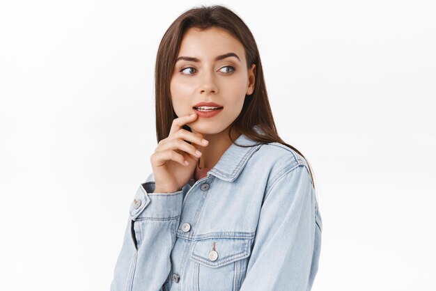 Pensativa sedutora garota elegante moderna na jaqueta jeans olhe ao redor contemplando algo tentador e interessante curiosamente olhando tocando lábio em pé fundo branco Copie o espaço