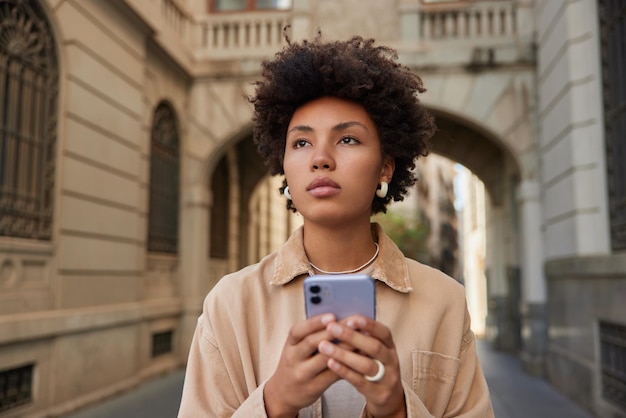 Pensativa turista de cabelos encaracolados usa aplicativo em smartphone passeia na cidade urbana durante férias solo concentrada na distância lê publicação de viagens se conecta à internet sem fio