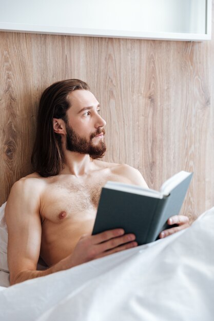 Pensativo barbudo jovem lendo livro e pensando na cama