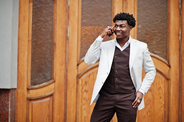 Pensativo e bonito cavalheiro afro-americano em roupas formais Modelo preto elegante homem de jaqueta branca falar no telefone
