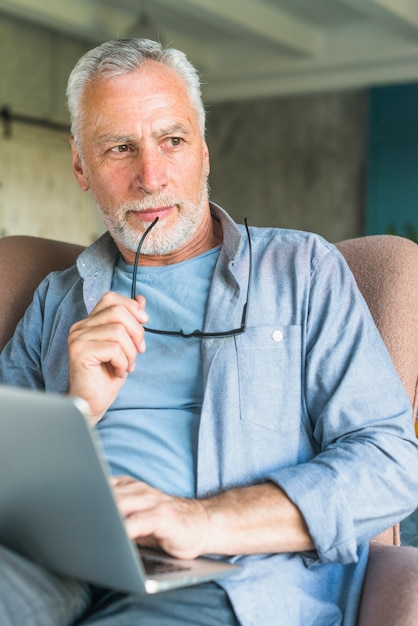 Pensativo, homem sênior, segurando, óculos, sentando, em, poltrona, com, laptop