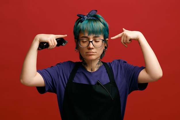 Pensativo jovem barbeiro feminino usando óculos uniformes e bandana segurando o aparador de cabelo fazendo gesto de brainstorming com os olhos fechados, isolados no fundo vermelho