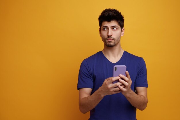 Pensativo jovem bonito segurando o celular olhando para o lado no fundo amarelo com espaço de cópia