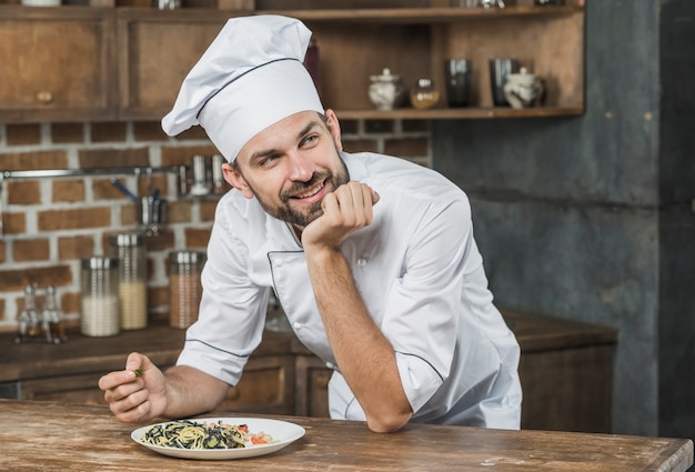 Pensativo sorridente chef masculino encostado no balcão com um prato na cozinha