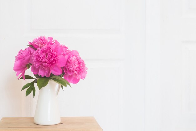 Peônias rosa em vaso esmaltado branco. Ramalhete das flores na tabela de madeira no interior branco de provence. Interior da casa com elementos de decoração