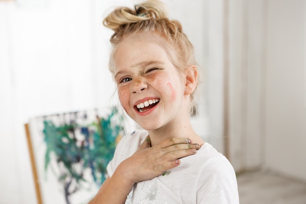 Pequena menina loira Europeia com rosto pintado, rindo e apertando os olhos na luz da manhã. Humor criativo e atmosfera alegre misturado com olhar brilhante de garoto vestindo camiseta branca.