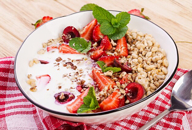 Pequeno-almoço saudável - granola, morangos, cereja, nozes e iogurte em uma tigela sobre uma mesa de madeira. Comida de conceito vegetariano.