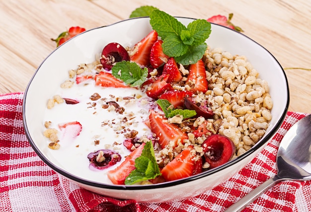 Pequeno-almoço saudável - granola, morangos, cereja, nozes e iogurte em uma tigela sobre uma mesa de madeira. Comida de conceito vegetariano.