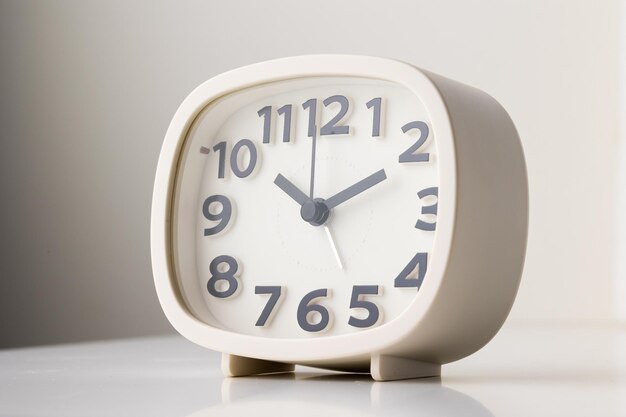 Pequeno despertador branco, números pretos, acertou a hora para 10h10, colocado sobre uma mesa branca.