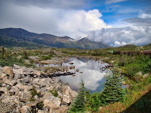 Pequeno lago cercado por montanhas e vegetação sob um céu azul nublado - perfeito para papéis de parede