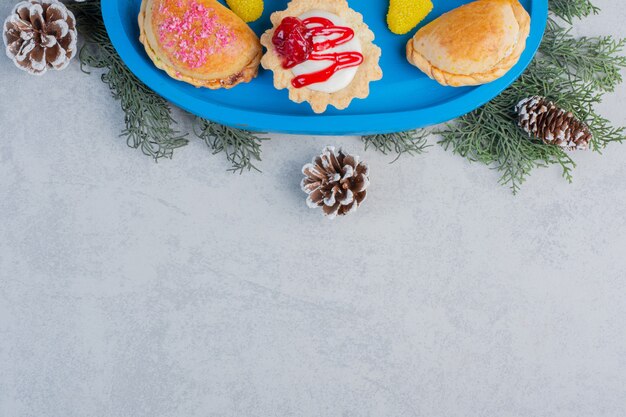 Pequenos pães, um cupcake e balas de geleia em uma bandeja azul decorada com folhas de pinheiro e cones em superfície de mármore