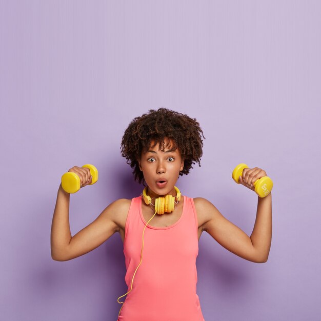 Perda de peso e conceito de exercício. Mulher de pele escura surpresa com cabelo encaracolado, levanta halteres, treina músculos, faz exercícios fáceis para bíceps, usa blusa rosa casual, usa fones de ouvido
