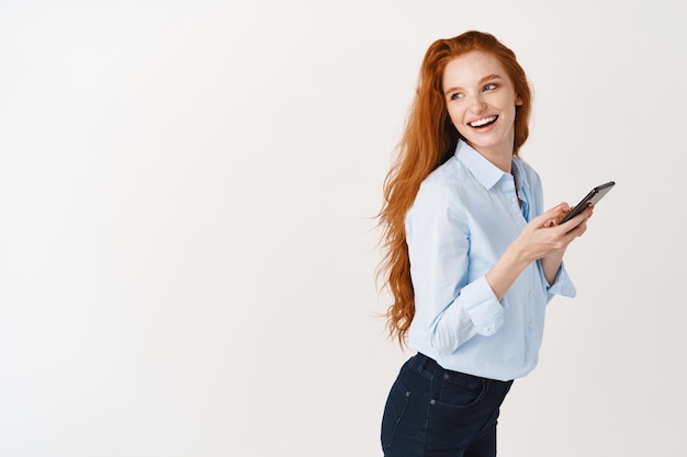 Perfil da bela gerente feminina com longos cabelos ruivos usando telefone celular, vire a cabeça para a esquerda e sorrindo para o espaço da cópia, em pé com o smartphone contra uma parede branca