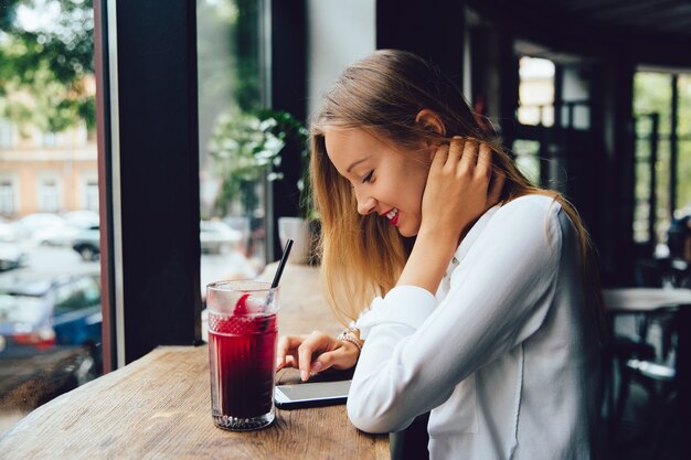 Perfil da mulher loura nova de sorriso, usando um smartphone, texting uma mensagem