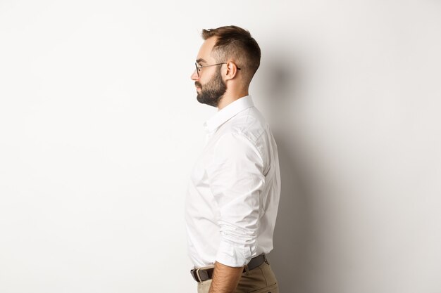 Perfil de jovem empresário em camisa de colarinho branco e calça bege, olhando para a esquerda, em pé contra o fundo do estúdio.