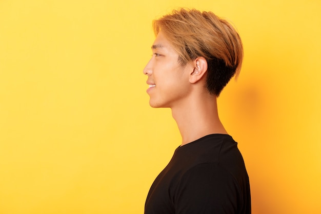 Perfil de um asiático bonito elegante com cabelo loiro, olhando para a esquerda e sorrindo, em pé sobre a parede amarela