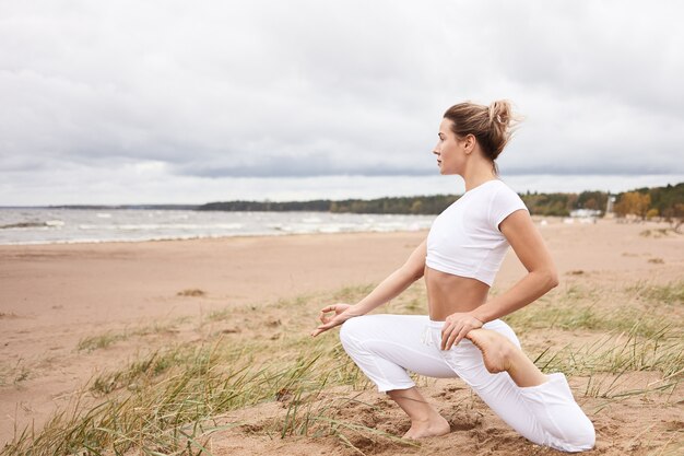 Perfil horizontal de uma bela jovem loira atlética malhando na praia, de frente para o mar, fazendo exercícios de aquecimento e alongamento durante a prática de ioga, sentada em Eka Pada Rajakapotasana
