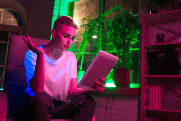 Perguntando. Retrato cinematográfico de mulher elegante no interior iluminado por néon. Tons de efeitos de cinema, cores neon brilhantes. Modelo caucasiano usando tablet em luzes coloridas dentro de casa. Cultura jovem.