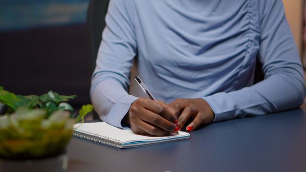 Perto da empresária africana, escrevendo a lista de tarefas para o projeto de negócios no caderno, sentado na mesa na sala de estar fazendo hora extra. Freelancer negro respeitando prazo de estudo tarde da noite