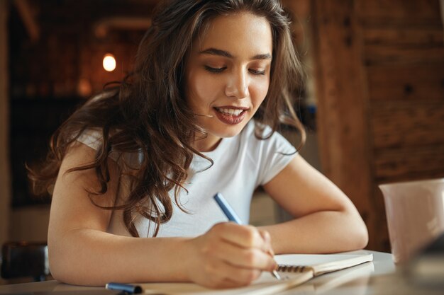 Perto da encantadora adolescente plus size com cabelos soltos ondulados, sentada na mesa com o caderno, caligrafia, desenhando ou fazendo esboços, tendo o olhar alegre. Conceito de criatividade, hobby e lazer