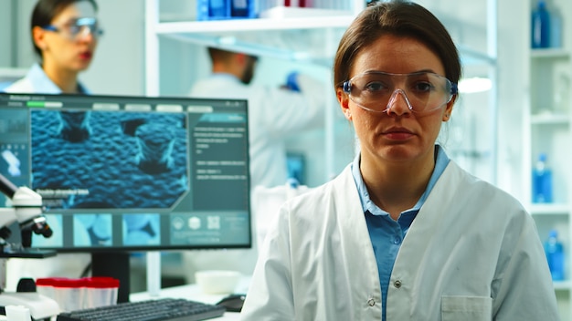 Perto da enfermeira cientista parecendo cansado para a câmera, sentado no moderno laboratório equipado, tarde da noite. equipe de especialistas examinando a evolução do vírus usando alta tecnologia para pesquisa e desenvolvimento de vacinas