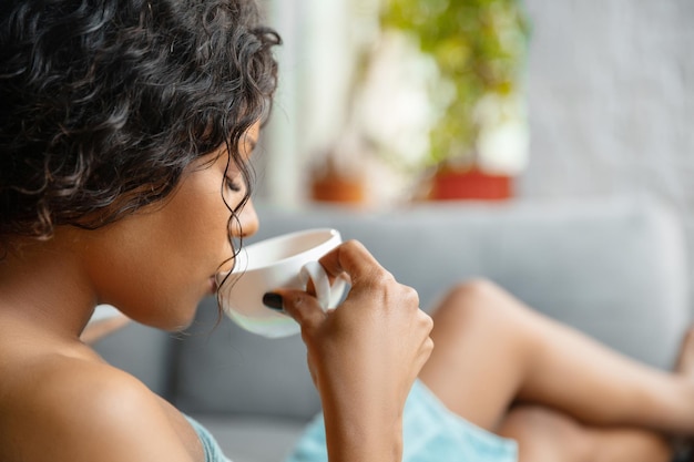 Perto da mulher afro-americana na toalha, fazendo sua rotina diária de beleza em casa. sentado no sofá, parece satisfeito, bebendo café e relaxando.