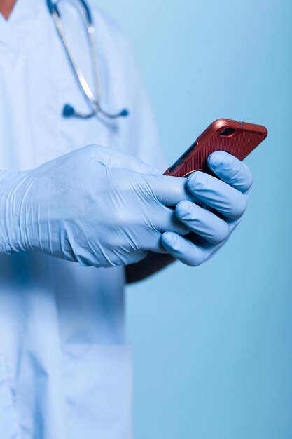 Perto da pessoa que trabalha como enfermeira, segurando o smartphone com tela de toque no estúdio. Médico assistente usando estetoscópio, uniforme azul e luvas enquanto usa o celular em um fundo isolado