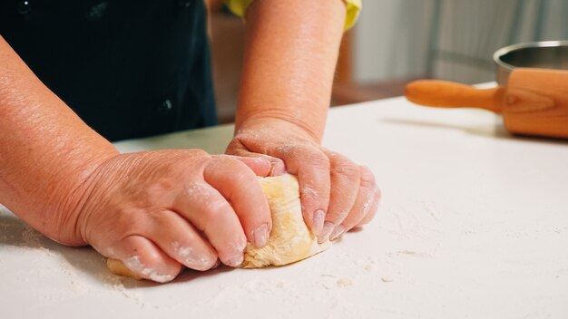 Perto das mãos da avó, amassando na mesa da cozinha em casa. Padeiro idoso aposentado com bonete misturando ingredientes com farinha de trigo peneirada amassando para assar pão e bolo tradicional.