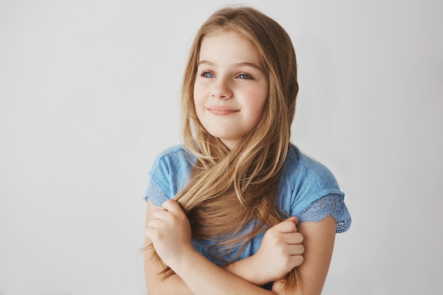 Perto de alegre menina loira de olhos azuis, segurando o cabelo com as mãos, olhando de lado com expressão feliz e relaxada.