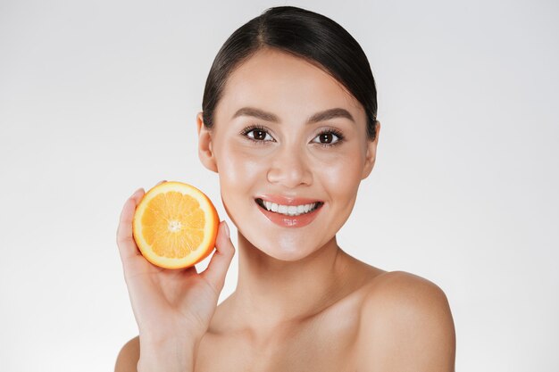 Perto de mulher satisfeita com a pele fresca saudável, segurando a laranja suculenta e sorrindo, isolado sobre o branco