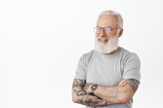 Perto de um cara bonito sênior com tatuagens, usando óculos, olhando para a esquerda e sorrindo feliz para a câmera, conversando com alguém, em pé com uma camiseta cinza contra um fundo branco