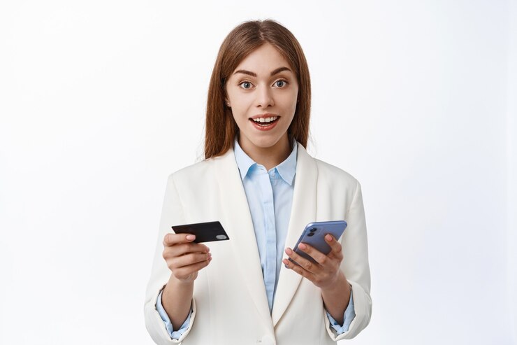 Foto grátis perto de uma jovem mulher de negócios em um terno fácil de pagar online com cartão de crédito de plástico e smartphone, fazer compras online, segurando um telefone móvel, parecendo espantado com a parede frontal branca