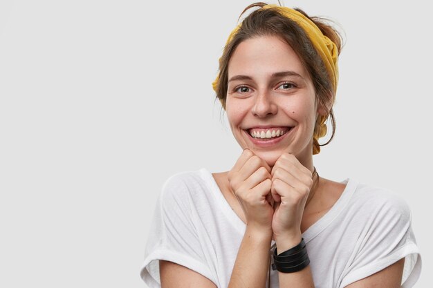 Perto de uma linda mulher sorridente com expressão facial alegre, mantém as mãos sob o queixo, olhando diretamente para a felicidade