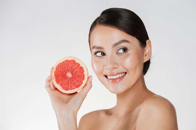 Perto de uma mulher feliz com a pele fresca saudável, segurando a toranja suculenta e olhando de lado com o sorriso, isolado sobre o branco