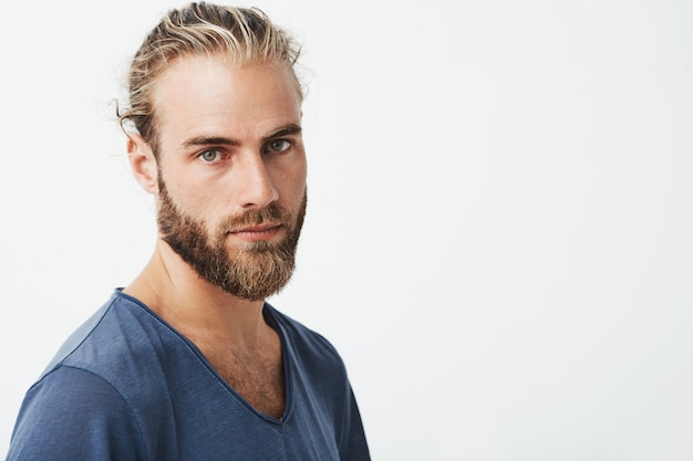 Perto do belo homem sueco com penteado elegante e barba na camiseta azul, olhando com expressão séria.