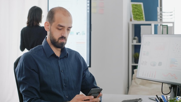 Perto do empresário usando smartphone com tela de toque no escritório. Homem com trabalho corporativo, segurando o telefone móvel, enquanto trabalhava com gráficos de dados no computador para projeto de negócios.