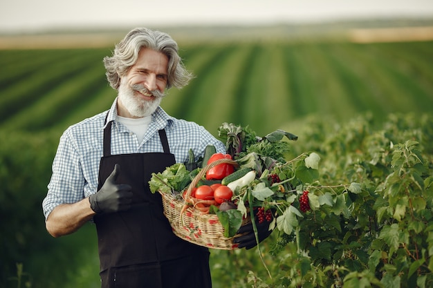 Perto do velho fazendeiro segurando uma cesta de legumes. O homem está parado no jardim. Sênior em um avental preto.