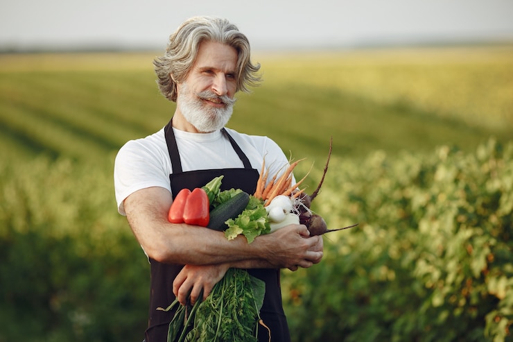 Foto grátis perto do velho fazendeiro segurando uma cesta de legumes. o homem está parado no jardim. sênior em um avental preto.