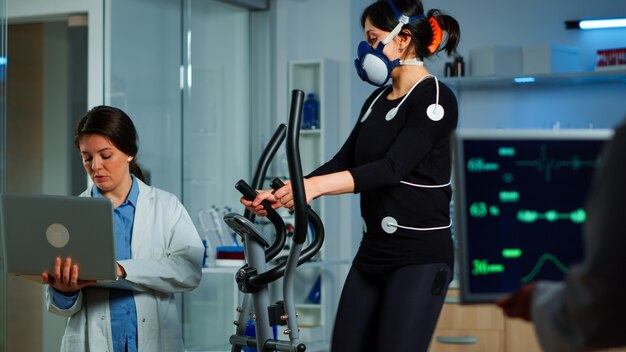 Pesquisador médico usando laptop enquanto mede a resistência do esportista usando sensores corporais, eletrodos e máscara para medir o ritmo cardíaco