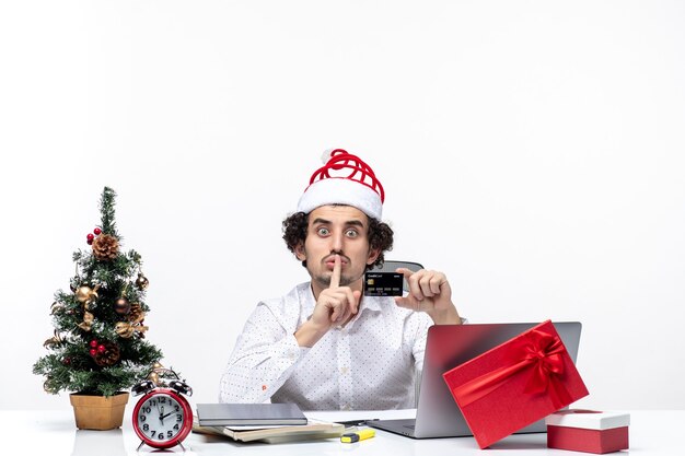 Pessoa de negócios com chapéu de Papai Noel e segurando o cartão do banco olhando surpreso fazendo gesto de silêncio no escritório