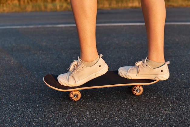 Pessoa desconhecida andando de skate na estrada de asfalto, pernas de mulher no longboard, mulher sem rosto usando tênis branco andando de skate.